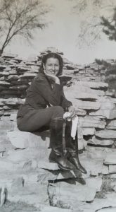 Young woman dressed in riding boots sits on rocks and poses for photo.